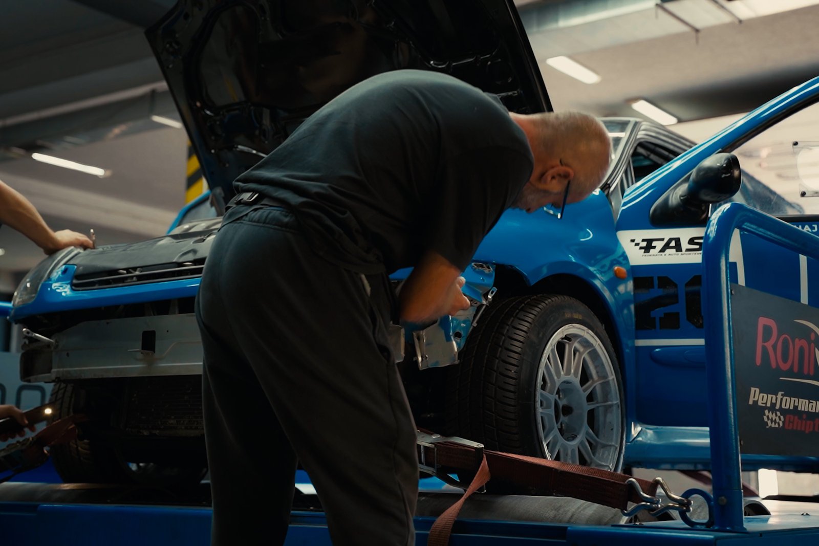 Color-graded still of a technician working on the blue car in the workshop.