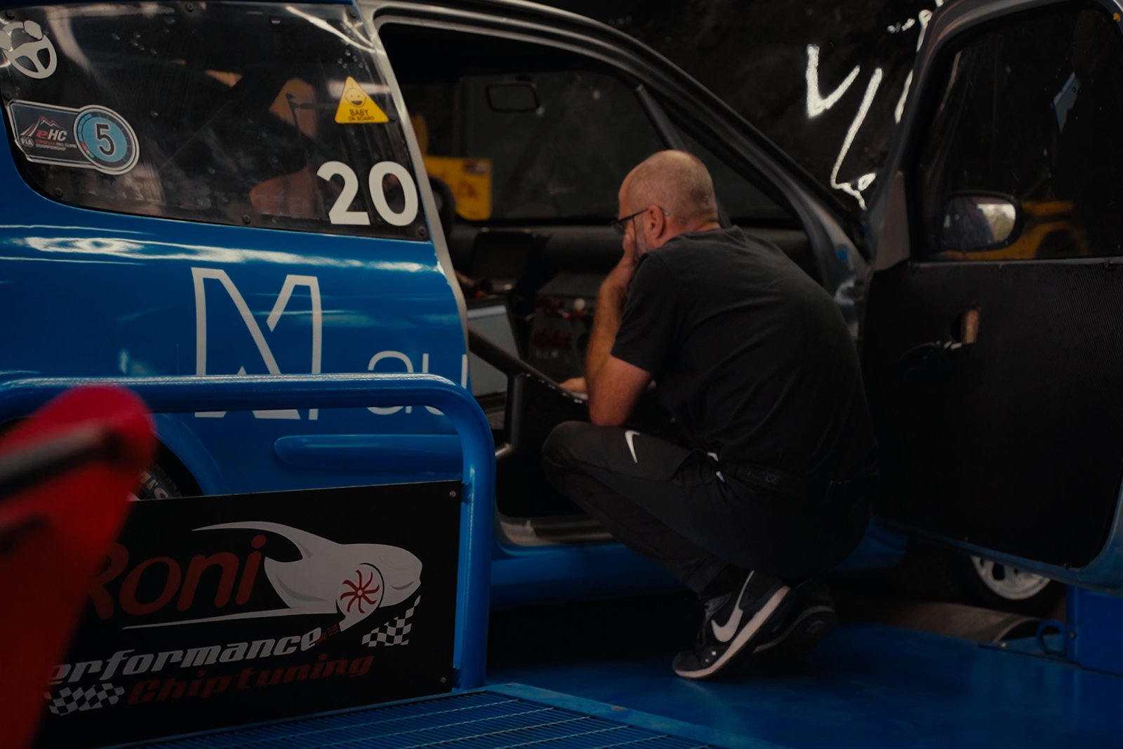 Color-graded still of a technician working beside the blue car at Roni Performance.